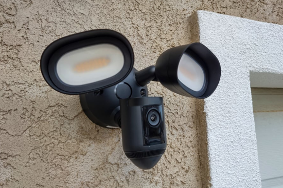 A black outdoor floodlight security camera mounted on a textured wall, featuring two bright LED lights and a camera in the center.