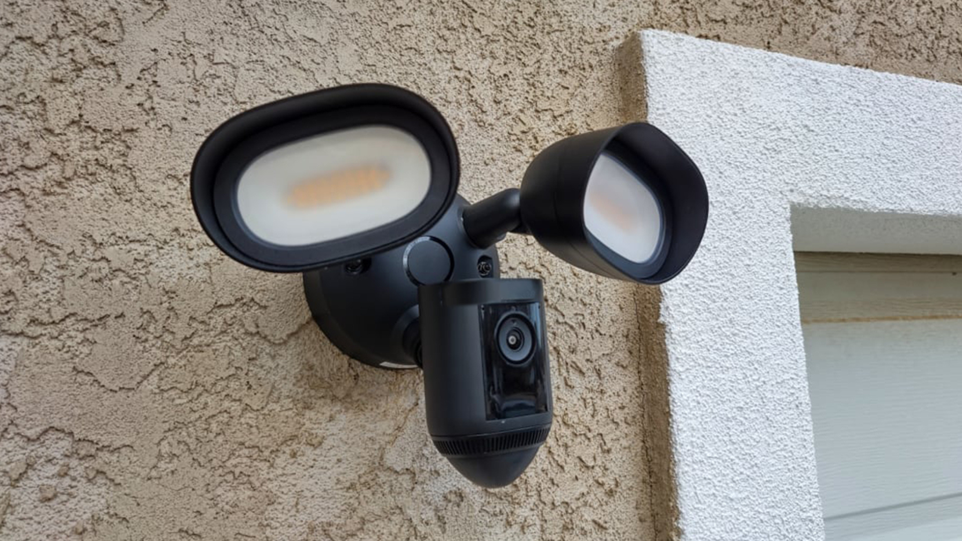 A black outdoor floodlight security camera mounted on a textured wall, featuring two bright LED lights and a camera in the center.