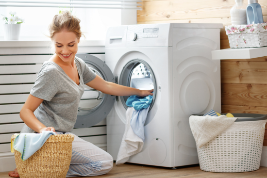 Person loading colorful clothes into a washing machine in a laundry room with detergent bottles and towels in the background