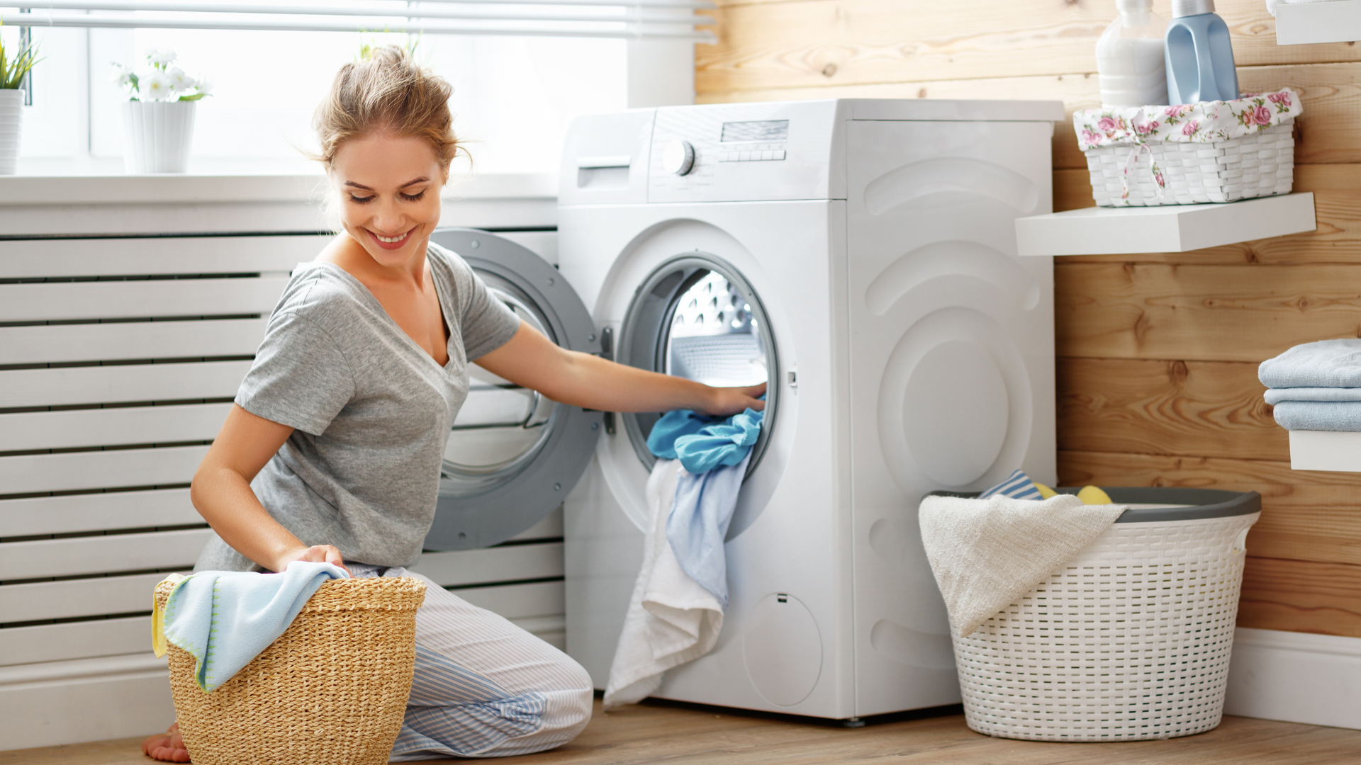 Person loading colorful clothes into a washing machine in a laundry room with detergent bottles and towels in the background