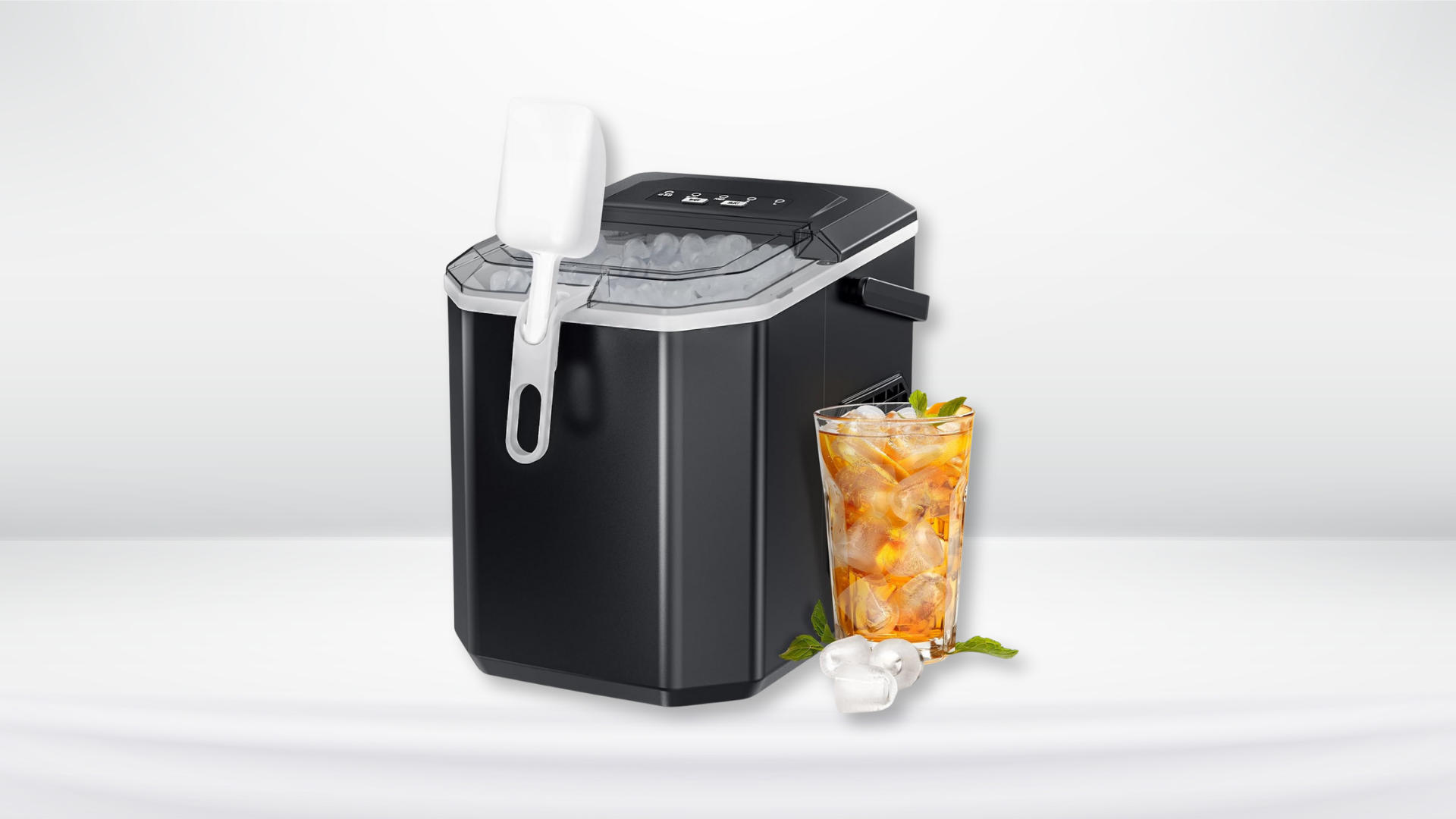 A black countertop ice maker with an ice scoop attached, alongside a glass of iced beverage garnished with mint leaves.