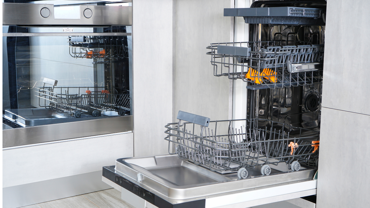 An open, empty dishwasher with stainless steel interior and racks extended, set in a modern kitchen next to a built-in oven.