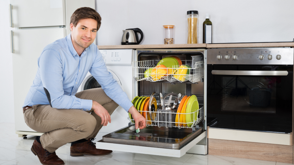 A man kneeling next to an open dishwasher filled with colorful dishes, in a modern kitchen with a washing machine and oven nearby.