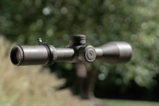 Close-up view of a rifle scope with adjustment dials, set against a blurred outdoor background, highlighting precision gear for hunting or target shooting.