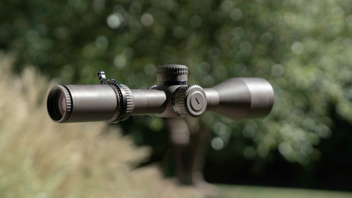 Close-up view of a rifle scope with adjustment dials, set against a blurred outdoor background, highlighting precision gear for hunting or target shooting.