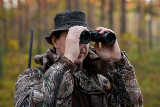 Hunter in camo jacket using binoculars in a forest, scanning the area during a wildlife or hunting trip.