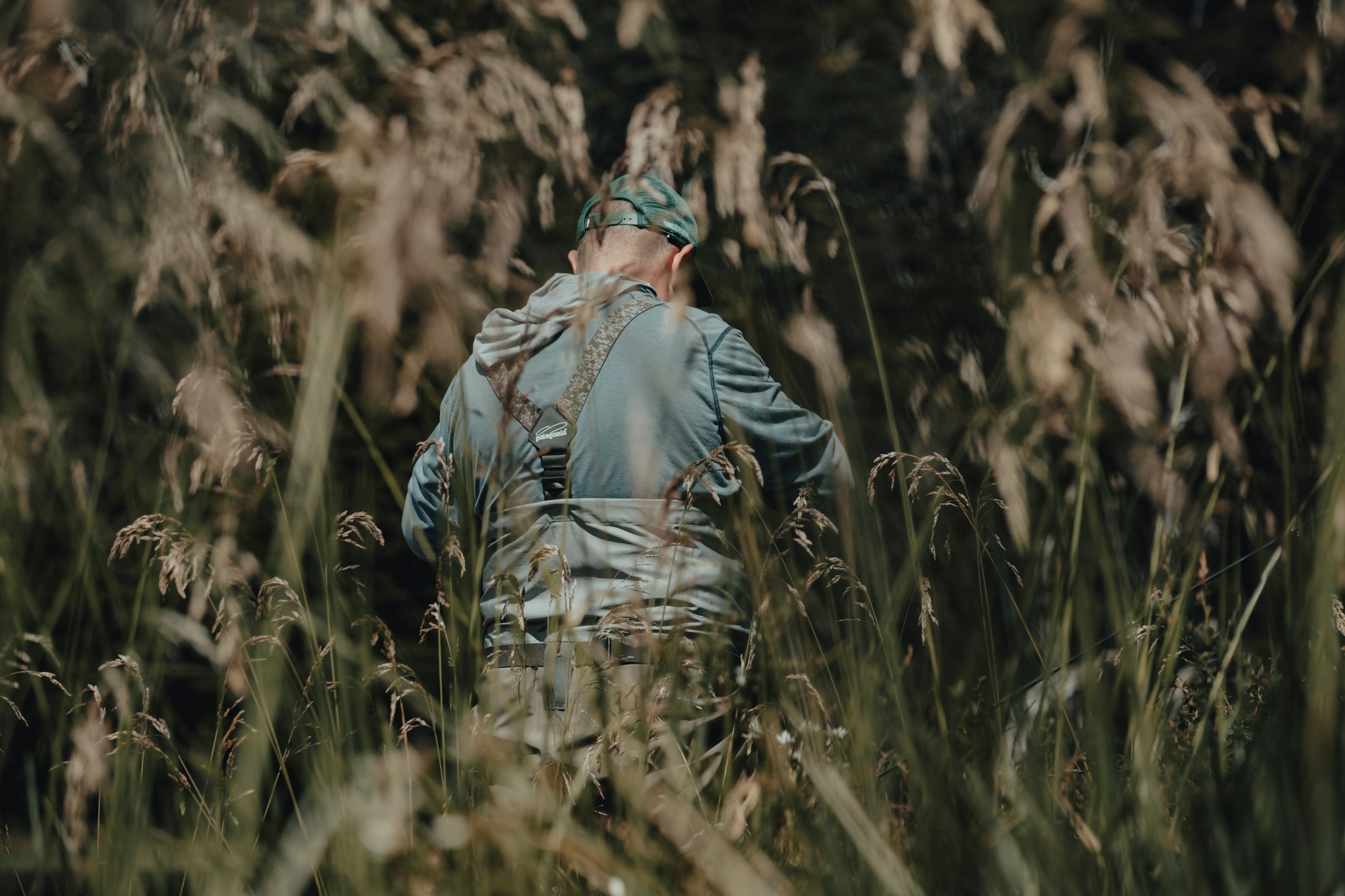 A man wearing outdoor gear and a cap walks through tall grass during a fishing or hunting trip in the woods.