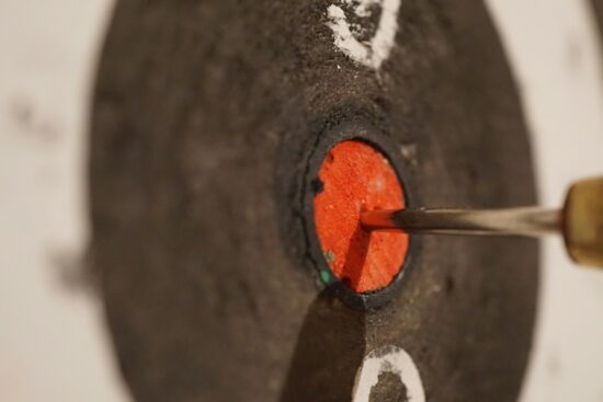 A close-up of a dart hitting the bullseye on a black and red target at an indoor range.