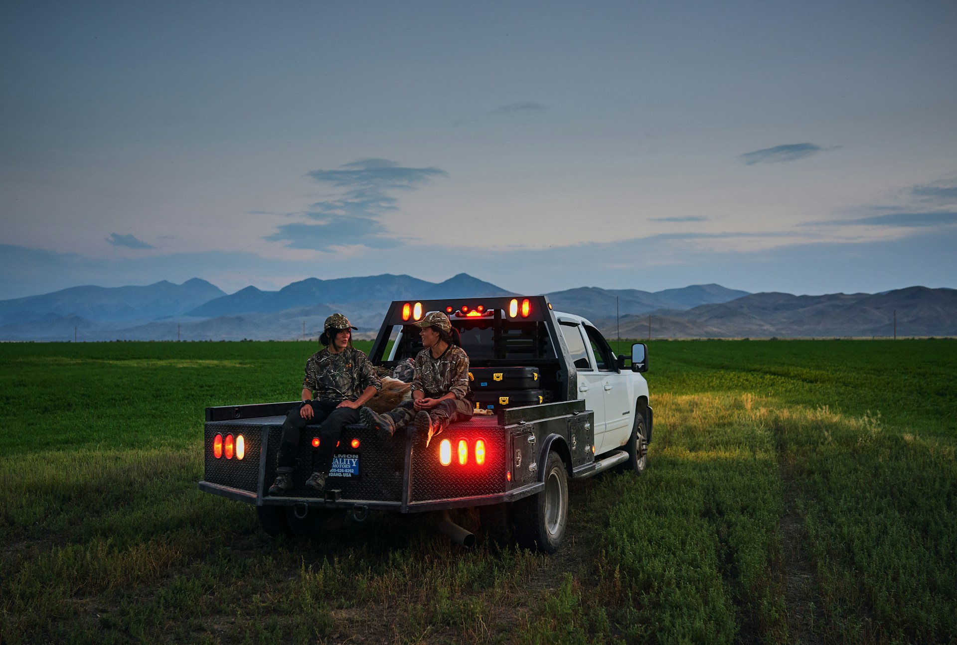 Two hunters in camo gear sitting on the back of a white pickup truck in an open field with mountains in the background at dusk.