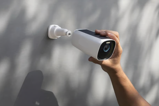 A person installing a white security camera onto a wall mount, preparing to secure it in place. The camera features a lens and solar panel on top.