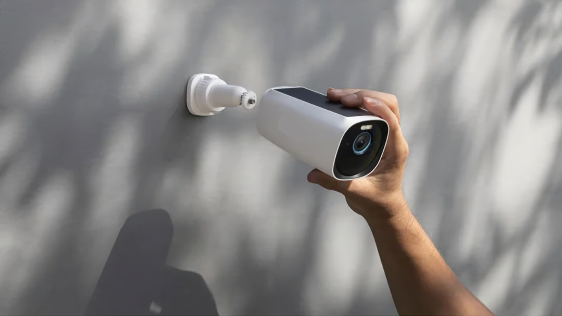 A person installing a white security camera onto a wall mount, preparing to secure it in place. The camera features a lens and solar panel on top.