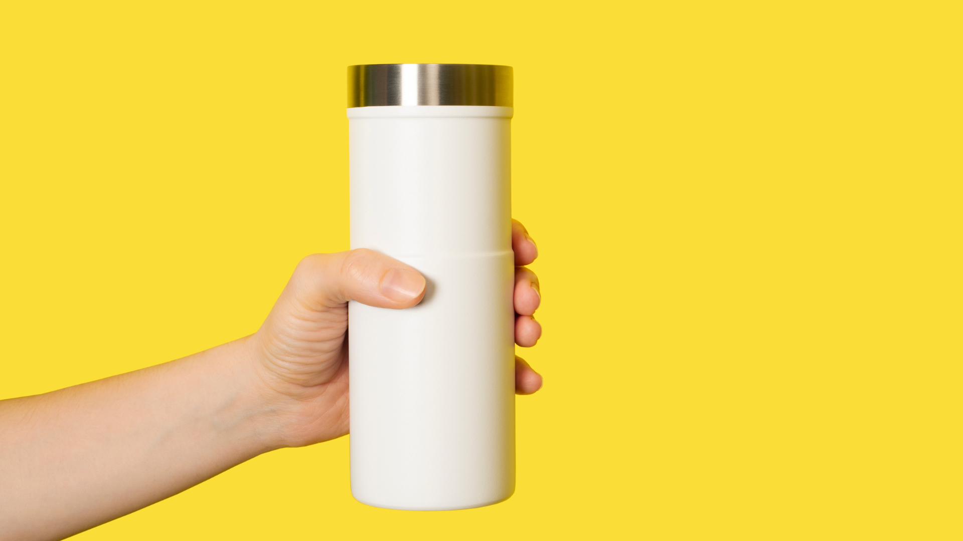 A hand holding a white tumbler with a silver lid, set against a bright yellow background.