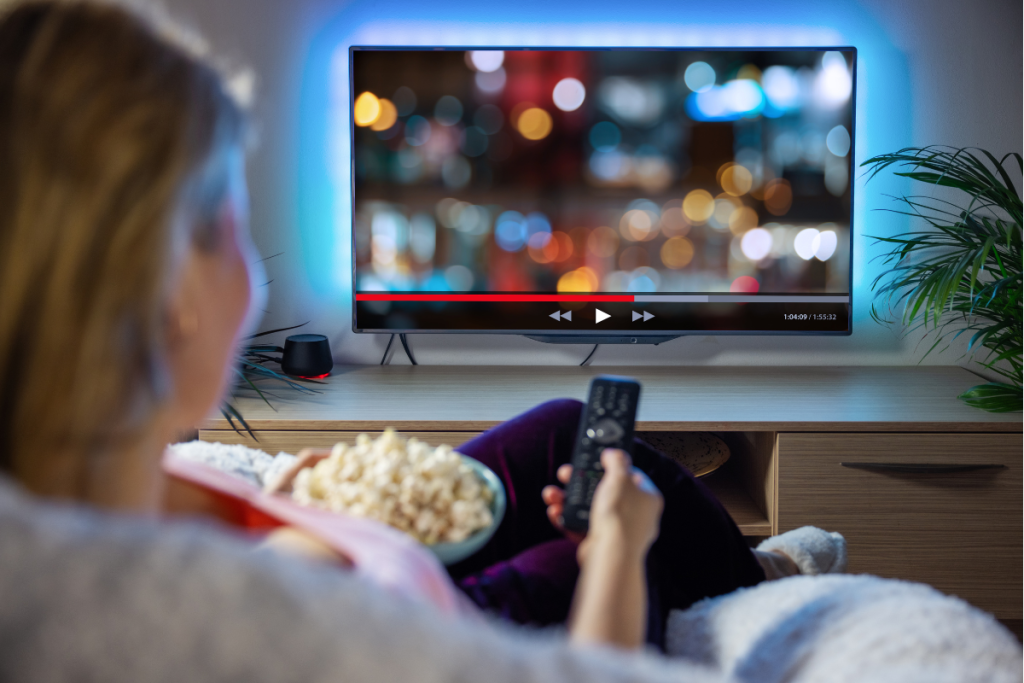 A person sitting on a couch holding a remote control and a bowl of popcorn while watching a movie on a flat-screen TV.