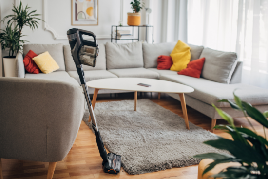 A cordless vacuum cleaner standing on a rug in a modern living room with a gray sectional sofa and colorful pillows.