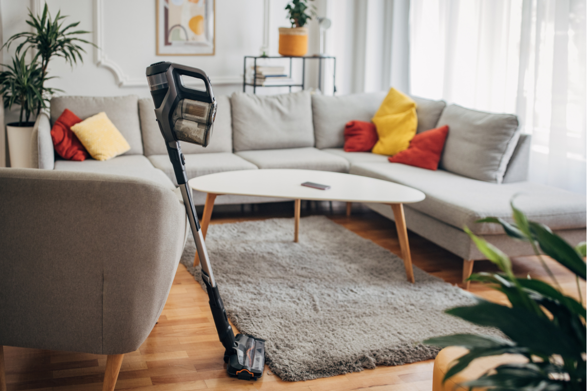 A cordless vacuum cleaner standing on a rug in a modern living room with a gray sectional sofa and colorful pillows.