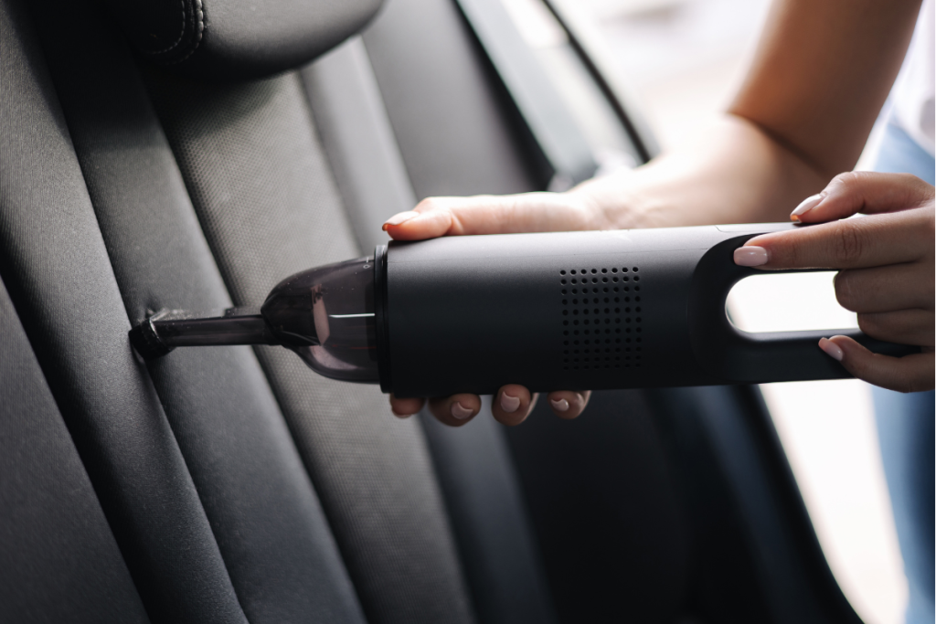 A person using a handheld vacuum cleaner to clean the crevice between the backrest and seat cushion of a car.