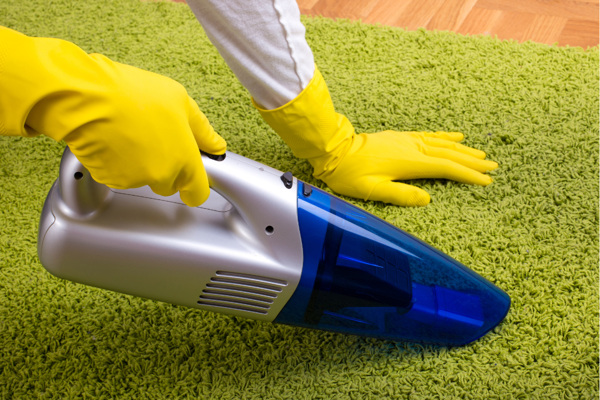 A person wearing yellow gloves using a handheld vacuum cleaner to clean a green carpet.