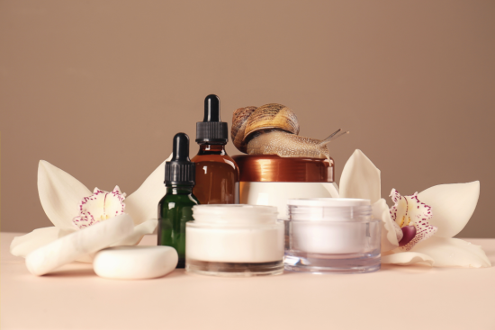 Skincare products, including dropper bottles and cream jars, arranged with white orchids and a snail on top of a container.