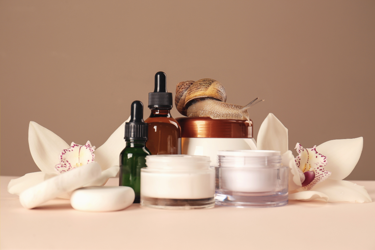 Skincare products, including dropper bottles and cream jars, arranged with white orchids and a snail on top of a container.