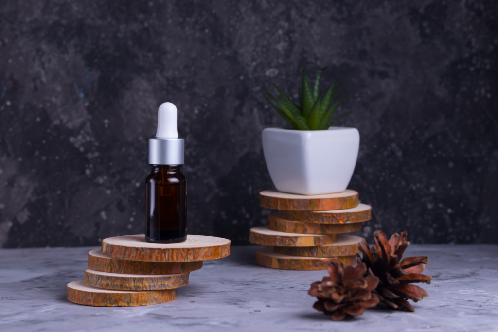 A dark amber dropper bottle placed on stacked wooden coasters with a small potted plant and pinecones in the background.