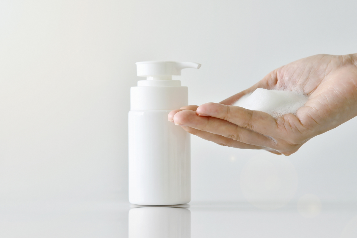 A hand holding a palmful of foam next to a white dispenser bottle against a minimalist white background.