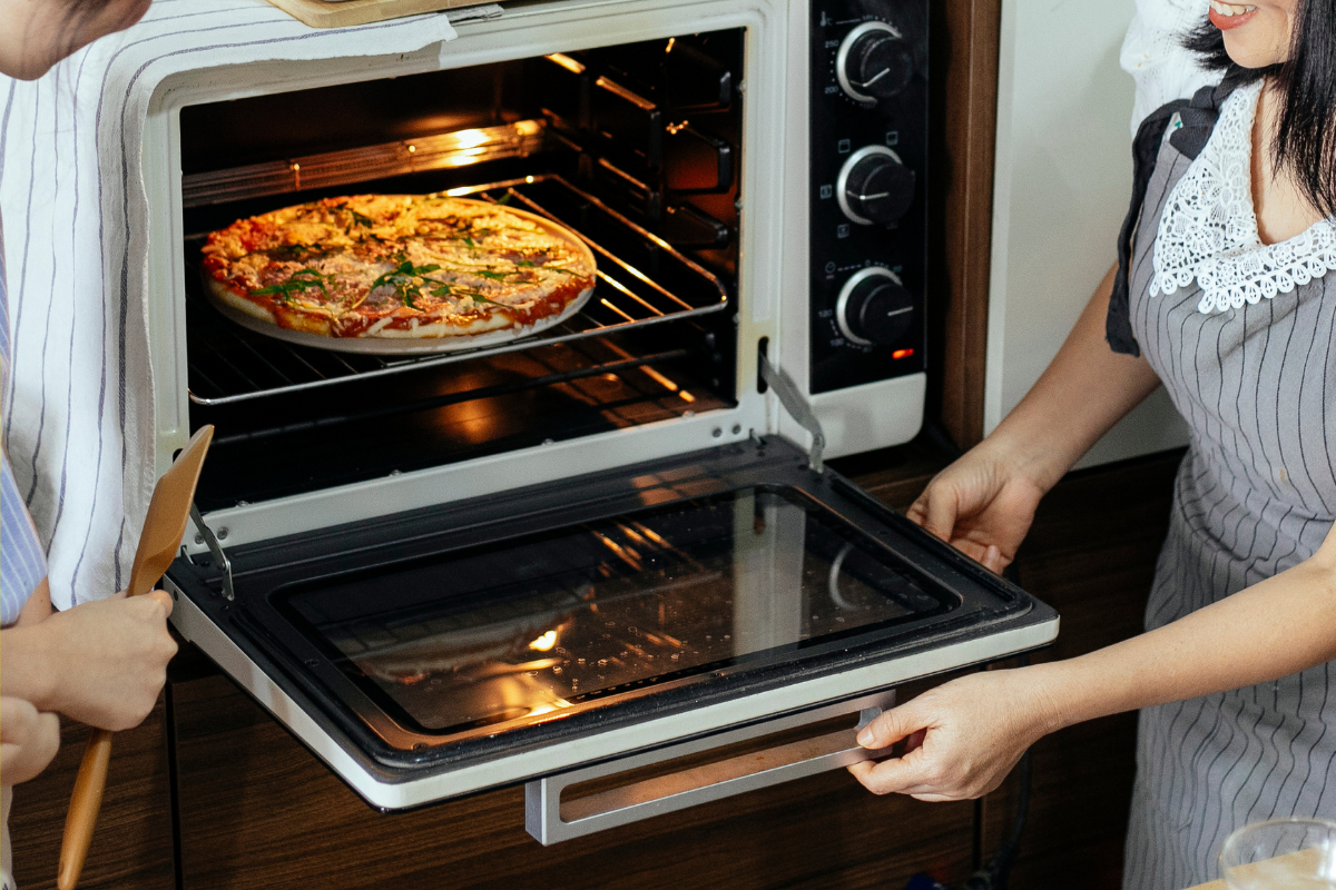 People taking a freshly baked pizza out of an oven in a kitchen setting.