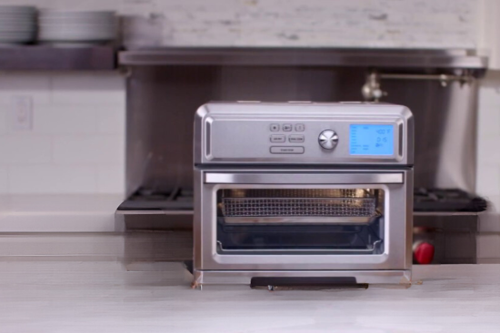 A modern stainless steel countertop oven with a digital display and control buttons, featuring a transparent door with a mesh tray inside.