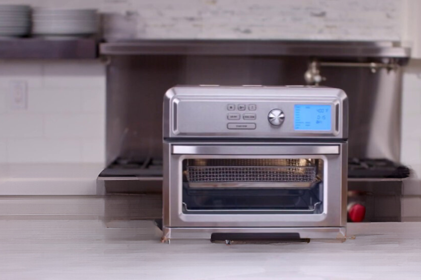 A modern stainless steel countertop oven with a digital display and control buttons, featuring a transparent door with a mesh tray inside.