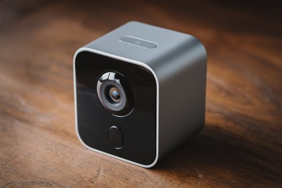 Compact, square-shaped pet camera with a glossy black front and metallic finish, placed on a wooden surface.