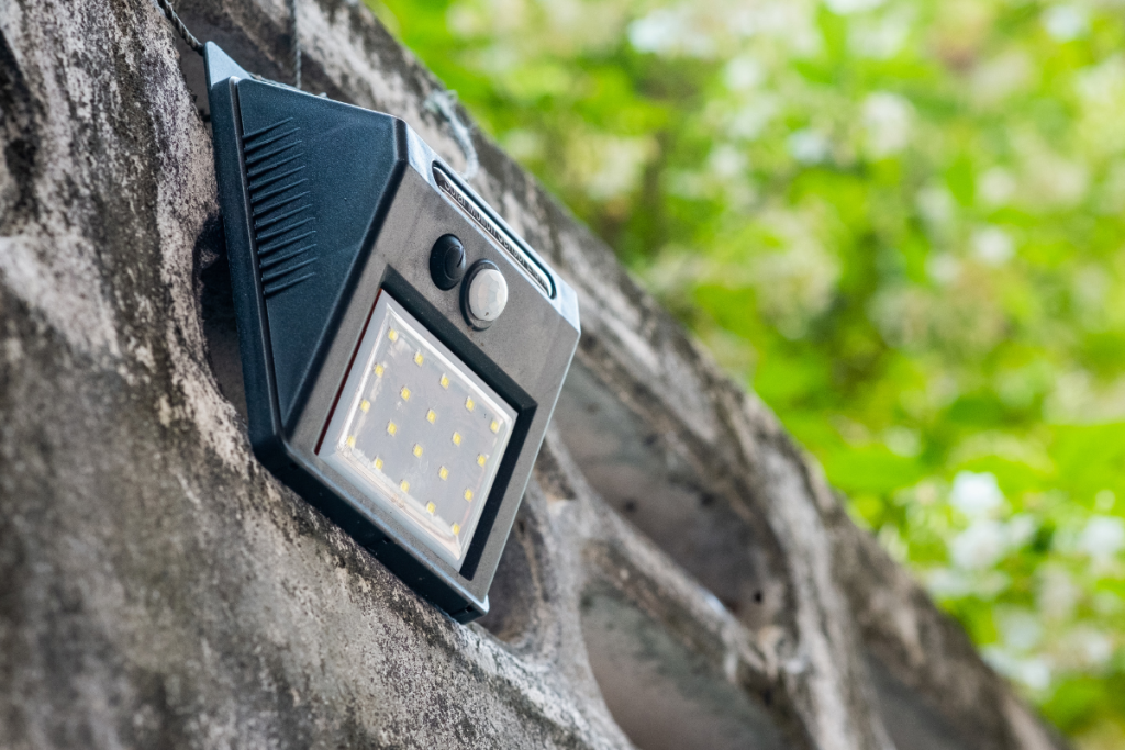 Black solar-powered motion sensor light mounted on a stone wall, with greenery in the background.