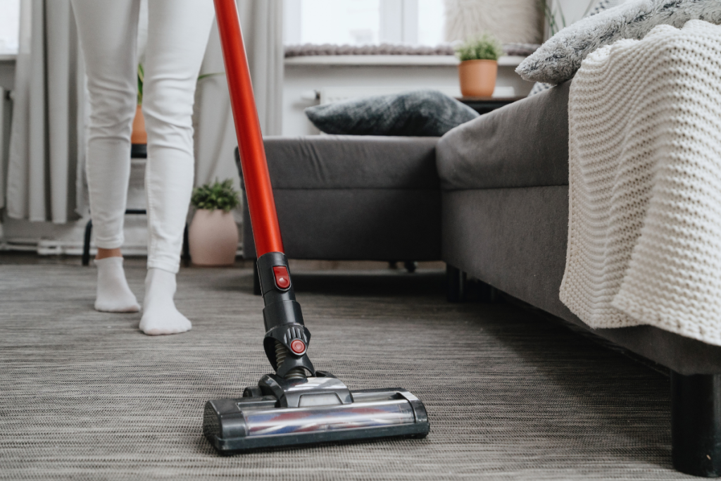 A person vacuuming a carpet near a gray sofa with a red cordless vacuum cleaner in a cozy, modern living room.