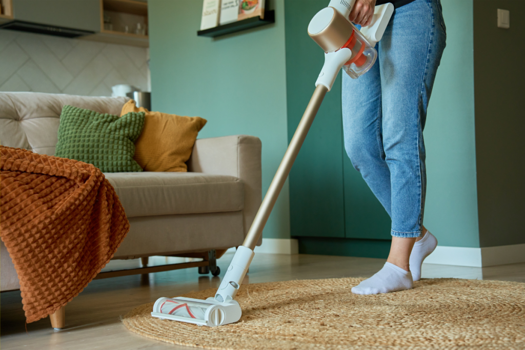 Person using a cordless stick vacuum cleaner on a round rug in a cozy living room with a beige sofa and colorful pillows.