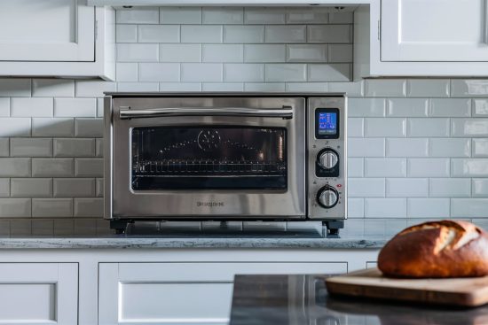 Stainless steel countertop oven on a marble countertop in a white kitchen, with a loaf of bread on a cutting board in the foreground.