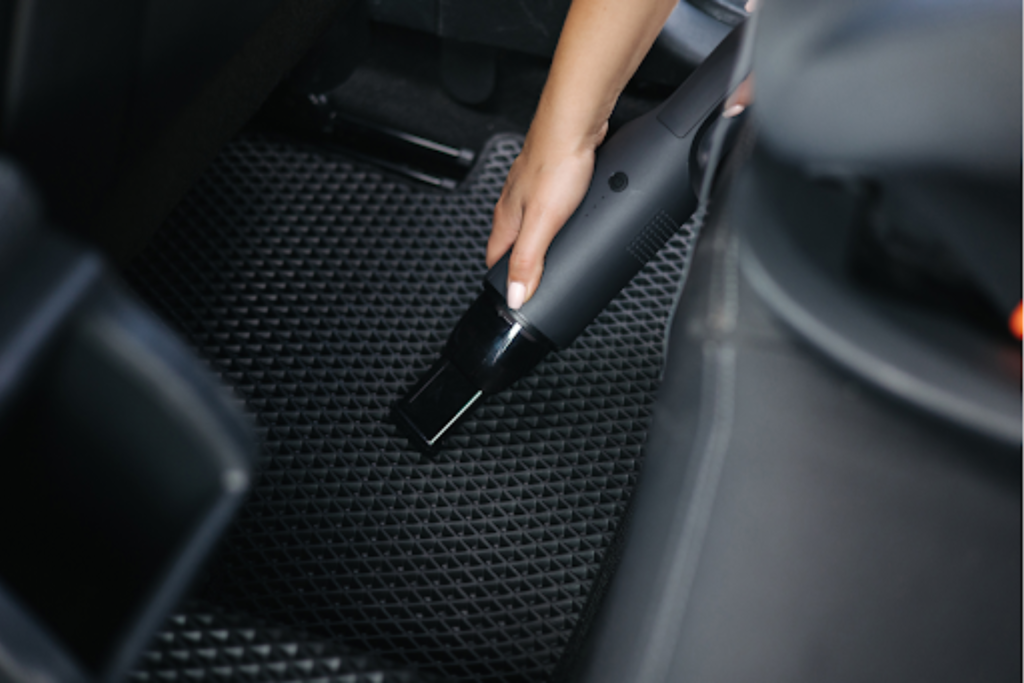 A person using a handheld vacuum cleaner to clean the floor mat of a car.