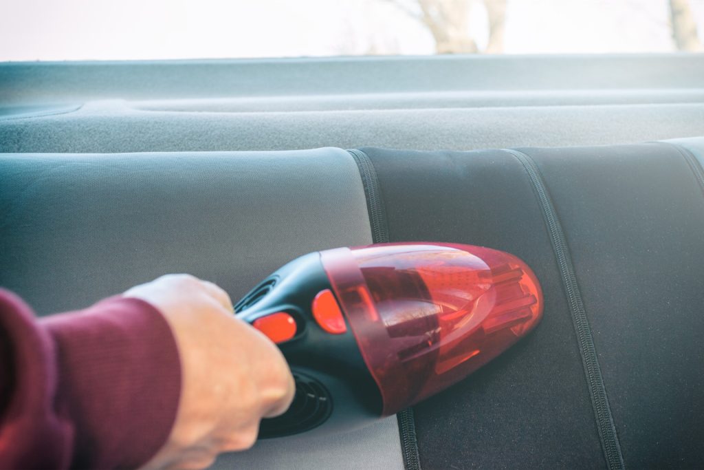 A person using a red handheld vacuum cleaner to clean the backseat of a car.