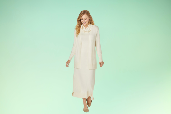 A smiling woman wearing a cream-colored long dress and scarf, walking gracefully against a soft green gradient background.