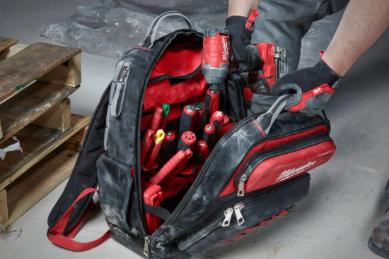 Construction worker organizing tools in a heavy-duty tool backpack with various compartments and red-handled tools.