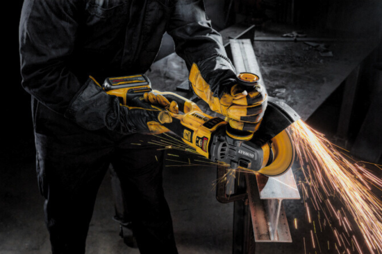 Worker using a yellow cordless angle grinder to cut a metal beam, producing bright sparks in a dimly lit workshop.