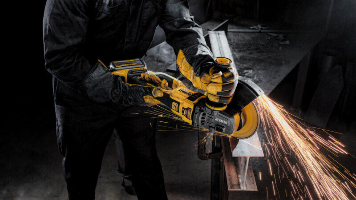 Worker using a yellow cordless angle grinder to cut a metal beam, producing bright sparks in a dimly lit workshop.