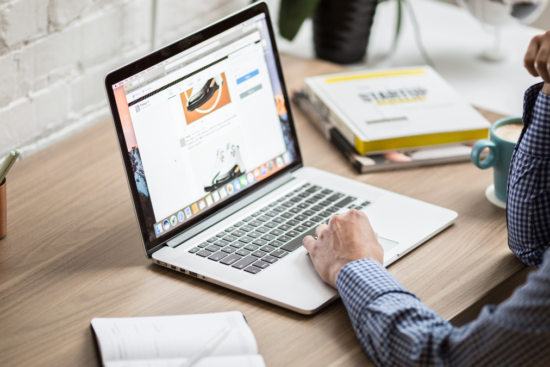 A person in a checkered shirt browsing on a laptop at a wooden desk, surrounded by notebooks, a cup, and books, with a bright and minimal workspace.