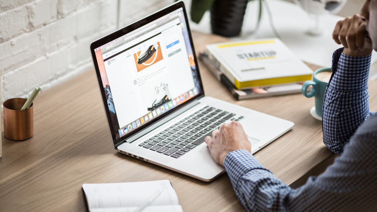 A person in a checkered shirt browsing on a laptop at a wooden desk, surrounded by notebooks, a cup, and books, with a bright and minimal workspace.
