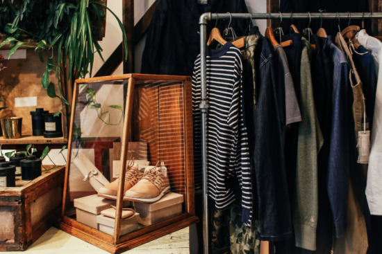 A cozy boutique setup featuring a clothing rack with striped and neutral-toned apparel, a wooden display case with shoes, and rustic decor elements like plants and vintage boxes.