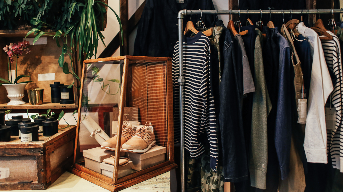 A cozy boutique setup featuring a clothing rack with striped and neutral-toned apparel, a wooden display case with shoes, and rustic decor elements like plants and vintage boxes.