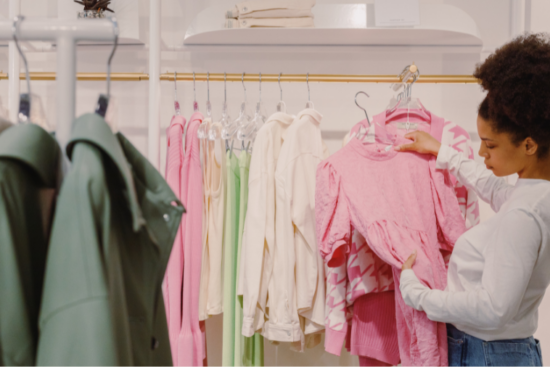 A woman shopping in a clothing store, browsing pink garments on a rack, with pastel and green clothes in the background.