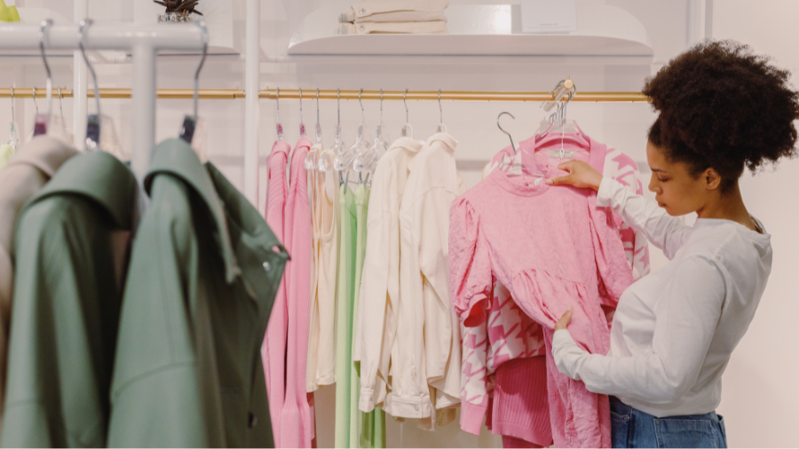 A woman shopping in a clothing store, browsing pink garments on a rack, with pastel and green clothes in the background.