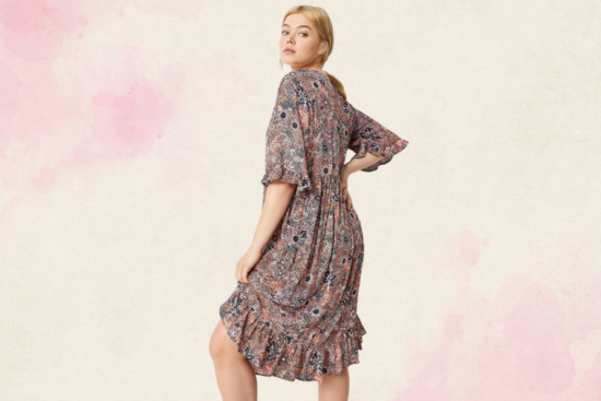 A woman wearing a patterned knee-length dress with ruffle details and elbow-length sleeves, posing against a light pink and beige background.