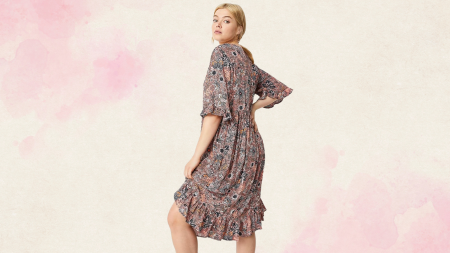 A woman wearing a patterned knee-length dress with ruffle details and elbow-length sleeves, posing against a light pink and beige background.