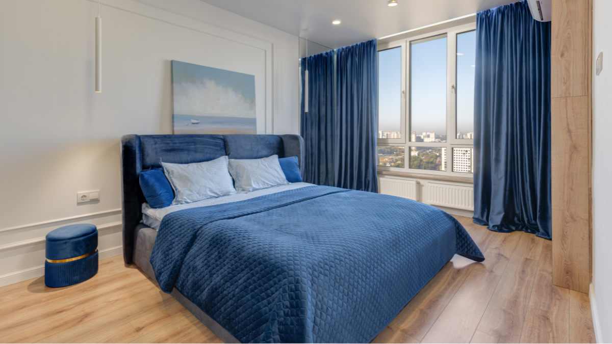 A modern bedroom featuring a blue upholstered bed, matching quilt, pillows, and curtains, with large windows overlooking a cityscape.