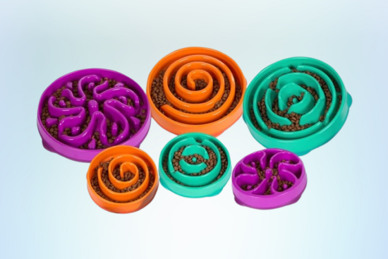 A set of colorful slow feeder dog bowls in purple, orange, and green, filled with kibble, displayed against a light gradient background.