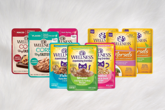 An assortment of Wellness cat food pouches, including CORE, Appetizing Entrées, and Morsels varieties, displayed against a soft white fur background.